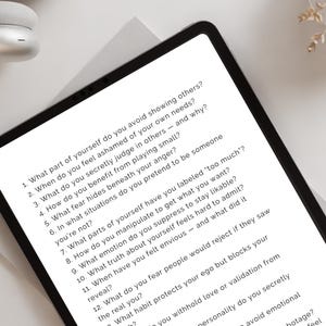 May include: A black tablet shows a list of self-reflection questions in white text. The tablet is on a white surface, with white headphones and a decorative plant in the background. The questions are about self-awareness.