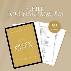 May include: A digital tablet and printed pages with journal prompts for grief. The tablet displays "Journal Prompts" in gold lettering. A gold circle with "365 prompts" is visible. The background is a light tan color.