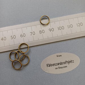 May include: Several small, brass-coloured metal rings. A ruler with centimetre markings is in the background. The rings are on a grey surface. The image also includes a label with the text "Ribbonzandcraftybitz on etsy.com".