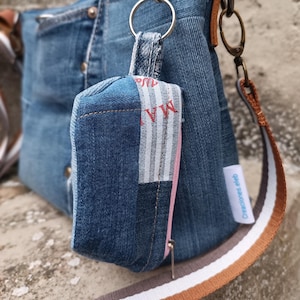 May include: A blue denim tote bag with a pink zipper and a brown and white striped strap. The bag has a small blue denim pouch attached to it with a silver key ring. The pouch has a white and grey striped fabric panel with the word "NEW" printed in red.