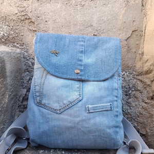Könnte beinhalten: Ein Jeans-Rucksack in verwaschenem Blau mit Klappenverschluss und verstellbaren Trägern. Der Rucksack hat eine Tasche und einen kleinen Libellen-Anhänger. Der Denim hat eine Tasche und einen kleinen Metall-Libellen-Anhänger.