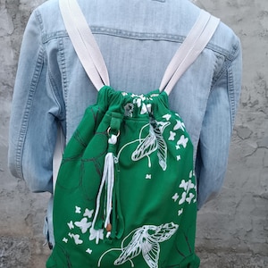 Könnte beinhalten: Grüner Rucksack mit Kordelzug und weißem Schmetterlings- und Blumendesign. Der Rucksack hat weiße Träger und eine weiße Quaste. Die Tasche ist aus Stoff und wird über den Schultern getragen.
