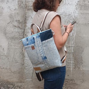 May include: A backpack constructed from repurposed blue denim fabric featuring a patchwork design with visible pockets, a top zipper closure, and brown and white striped webbing straps. The bag includes a small rectangular brown leather-like brand patch.