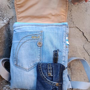 Può includere: Uno zaino in denim blu chiaro con chiusura a patta e una piccola tasca in denim attaccata. Lo zaino presenta un interno beige chiaro e una chiusura con bottone. La parola "Killah" è visibile sul denim.