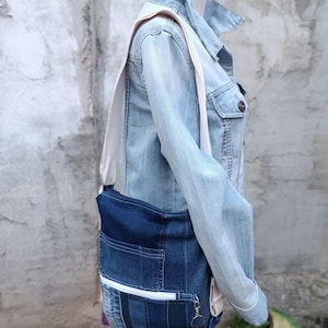 Puede incluir: Un bolso de hombro de mezclilla azul oscuro con una correa de color claro y cierre de cremallera. El bolso tiene dos bolsillos delanteros y un llavero. El bolso se lleva sobre una chaqueta vaquera azul claro.