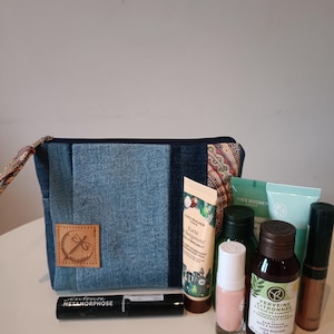 Puede incluir: Un neceser de mezclilla con un parche de cuero marrón y una variedad de productos de belleza. Los productos incluyen crema de manos, esmalte de uñas, rímel y varias botellas de productos para el cuidado de la piel. La bolsa está sobre una superficie blanca.