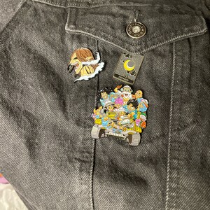 May include: A black denim jacket with three enamel pins. The pins are of a character from the anime Attack on Titan, a tarot card with a crescent moon, and a group of characters from the anime Dragon Ball Z.