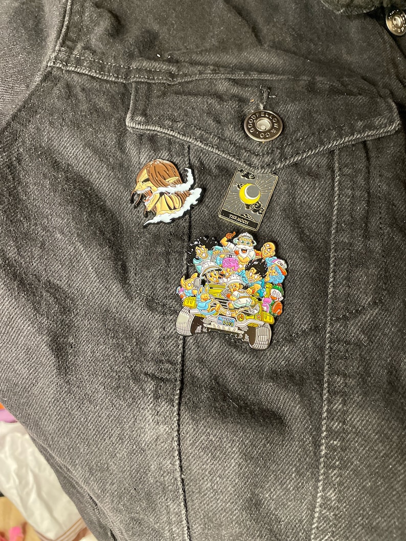 May include: A black denim jacket with three enamel pins. The pins are of a character from the anime Attack on Titan, a tarot card with a crescent moon, and a group of characters from the anime Dragon Ball Z.