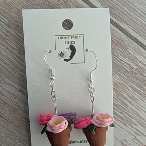 May include: Handmade earrings featuring miniature flower pots with pink and purple flowers. The earrings are attached to silver-colored fish hook ear wires. The earrings are displayed on a white card with the text "FRONT PAIGE FINDS" and a website address.