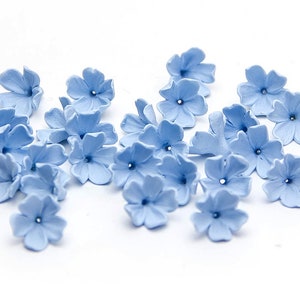 Puede incluir: Una colección de flores de papel azul claro, esparcidas sobre un fondo blanco. Las flores tienen cinco pétalos cada una y un pequeño agujero en el centro.
