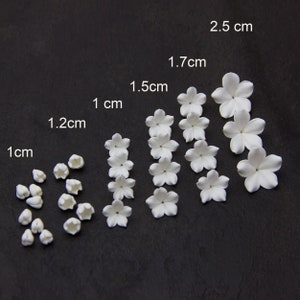 Pode incluir: Uma coleção de pequenas flores e botões brancos artificiais de vários tamanhos, variando de 1 cm a 2,5 cm. As flores são dispostas sobre uma superfície escura, mostrando seus detalhes e formas delicadas.