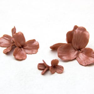 Puede incluir: Tres adornos individuales con forma de flor en un color rosa polvoriento apagado. Cada pieza presenta un punto central con tres pétalos redondeados distintos que irradian hacia afuera, mostrando una textura suave y mate y un tono sólido.