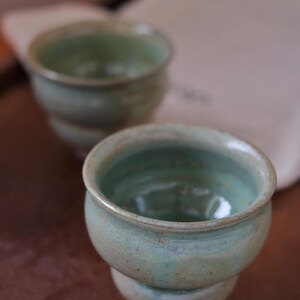 Può includere: Due tazze in ceramica verde chiaro fatte a mano con una superficie strutturata. Le tazze hanno una forma arrotondata e un bordo leggermente svasato. L'interno delle tazze ha una tonalità verde più scura. Le tazze sono su una superficie marrone.