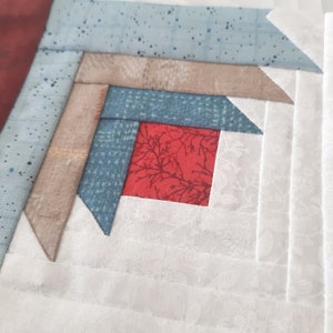 May include: A close-up of a quilt block with a red center square surrounded by layers of blue, beige, and white fabric. The fabric has a subtle pattern of small white dots.