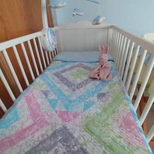 Puede incluir: Una cuna blanca con un edredón colorido con patrones geométricos rosas, azules y verdes. Un peluche de conejito rosa está sobre el edredón. Un móvil con adornos en forma de nube cuelga encima.