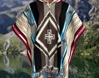 May include: A hooded poncho with a geometric pattern and a cross design. The poncho features a color palette of brown, black, white, blue, and red. It has a fringed hem and a zipper closure at the neckline. The background shows a mountain range and a lake.