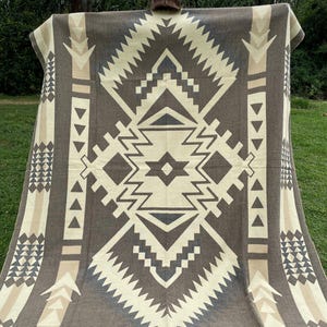 Puede incluir: Una manta marrón y crema con un patrón geométrico. La manta presenta una forma de diamante central con diseños en zigzag y triangulares. La paleta de colores incluye tonos de marrón, crema y beige. La manta se muestra al aire libre.