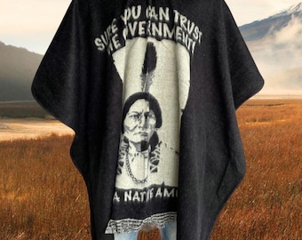 May include: Black hooded poncho with a Native American portrait and the text "SURE YOU CAN TRUST THE GOVERNMENT! A NATIVE AMERICAN". The poncho has a button closure and a fringed hem. The background shows a field and mountains.