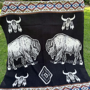 Puede incluir: Manta negra con un diseño nativo americano. La manta presenta ilustraciones de bisontes y calaveras blancas. El borde tiene un patrón geométrico en tonos marrones, azules y rojos. La manta se exhibe al aire libre sobre hierba verde.