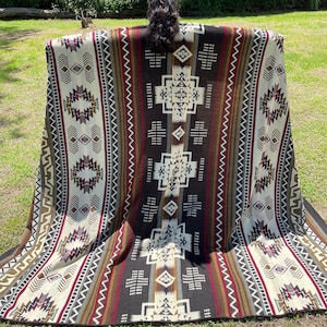 Peut inclure: Une grande couverture rectangulaire avec un motif géométrique du Sud-Ouest. La couverture présente un panneau central marron foncé avec des motifs géométriques blancs et beiges, flanqué de sections à motifs crème et marron. La couverture est exposée à l'extérieur sur une surface herbeuse.