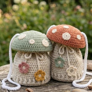 Op de afbeelding: Twee paddenstoelvormige gehaakte tassen met witte bandjes. Eén tas is lichtgroen met witte stippen, de andere is oranje met witte stippen. Beide hebben bloemenapplicaties en trekkoordsluitingen. Ze liggen op een houten tafel.