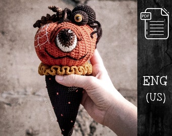 Puede incluir: Cono de helado de calabaza tejido a crochet con una araña en la parte superior. La calabaza es naranja con una boca y un ojo cosidos negros. El cono es marrón con cuentas negras y rojas. La araña es negra con un ojo amarillo.