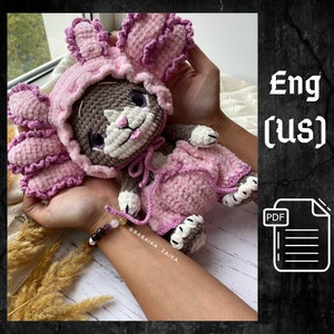 May include: A crocheted pink bunny rabbit toy with brown fur, white accents, and pink ears. The bunny is wearing a pink crocheted dress and a pink crocheted hat. The toy is being held in a person's hands.