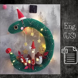 May include: A green crocheted wreath shaped like a crescent moon with a red Santa hat on top. The wreath is decorated with small crocheted houses, gold stars, and white lights. The text "Eng (US)" and "PDF" are visible in the image.