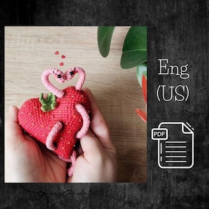 May include: A crocheted strawberry heart with two pink worms wrapped around it. The worms have black eyes and are holding each other in a heart shape. The strawberry is red with green leaves. The image is labeled "Eng (US) PDF".