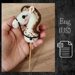 May include: A handcrafted, white crochet horse head with a brown mane and gold accents. The horse has a black bridle and is embellished with sequins. It is attached to a wooden stick, possibly for a craft project or toy.