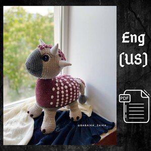 Puede incluir: Un juguete de dinosaurio Triceratops tejido a mano en tonos grises, beige y burdeos con lunares blancos. El juguete tiene cuernos y cola. La imagen incluye el texto "Eng (us)" y un icono PDF.