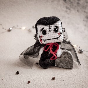 May include: A crocheted vampire doll with a black cape and a red heart. The doll has a white face with black eyes and a red mouth.