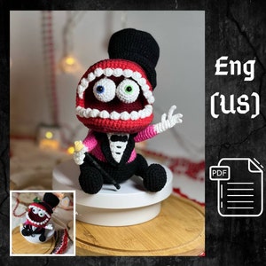 Puede incluir: Juguete amigurumi de crochet de una boca roja con dientes blancos, que lleva un sombrero de copa negro y un esmoquin rosa. El juguete tiene ojos verdes y azules y sostiene un bastón negro. El juguete está sentado en un pedestal blanco.