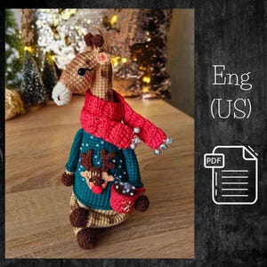 Puede incluir: Una jirafa de ganchillo hecha a mano, con una bufanda roja y un suéter verde azulado con un diseño de reno. La jirafa tiene pelaje marrón y beige, y el suéter tiene un pequeño bolsillo. La imagen también incluye un icono PDF y el texto "Eng (US)".