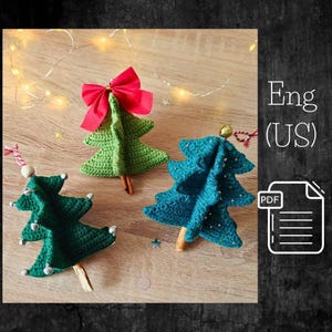 May include: Three handmade crochet Christmas tree ornaments. One green tree has a red bow, another is teal with gold accents, and the third is dark green with silver bells. Each tree has a wooden trunk.