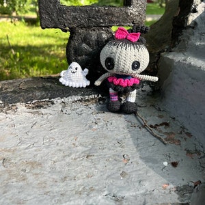 The crochet doll with its ghost companion standing on a step in a cozy outdoor setting. The black, gray, and pink color combination makes it a striking addition to any Halloween decor.