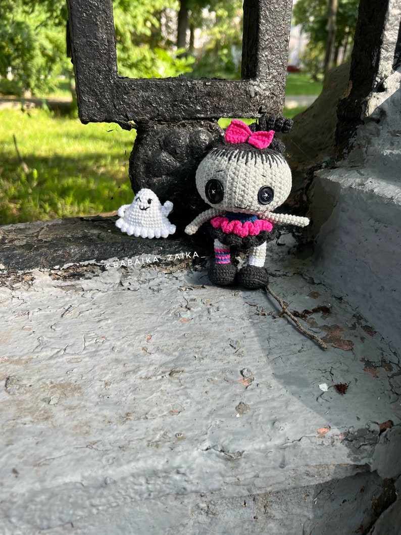 The crochet doll with its ghost companion standing on a step in a cozy outdoor setting. The black, gray, and pink color combination makes it a striking addition to any Halloween decor.