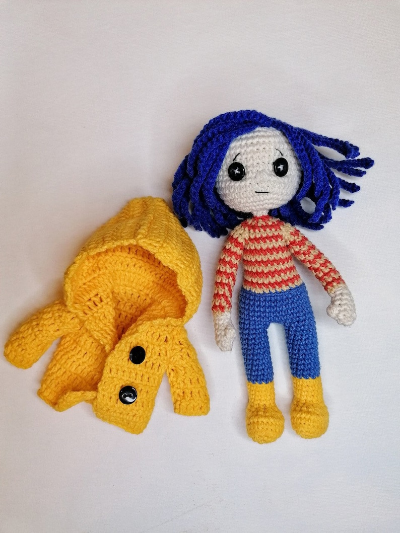 A display of the Coraline doll with her signature blue yarn hair, yellow coat, and detailed accessories, perfect for Halloween or collectible handmade decor.