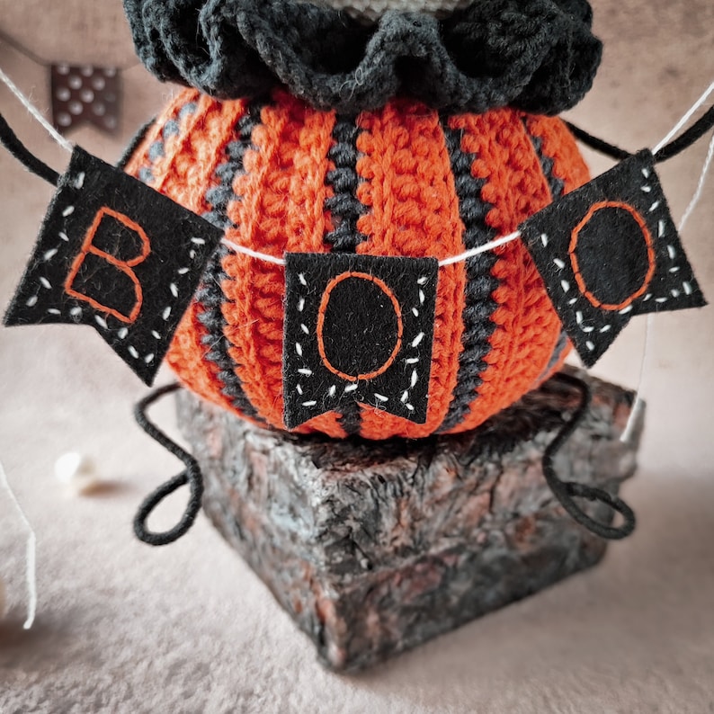 The crochet Jack pumpkin figure posed next to small Halloween decorations, with a spooky backdrop featuring a garland. The doll’s detailed crochet texture and handcrafted stitches are visible.