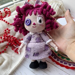 The crochet doll standing upright, with a colorful, patterned cloth underneath and white decorative beads.