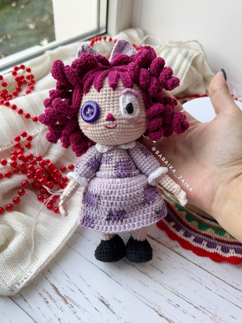 The crochet doll standing upright, with a colorful, patterned cloth underneath and white decorative beads.