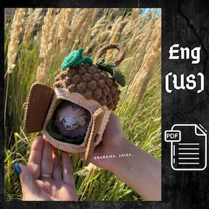 May include: A crocheted hedgehog in a brown and beige house with a green leafy roof. The house has two open doors and the hedgehog is inside. The house is being held in a person's hand. The text "Eng (US)" and "PDF" are visible in the image.