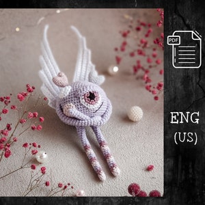 May include: A crocheted purple monster with a single eye, pink wings, and a pink heart on its head. The monster is sitting with its legs crossed and its arms wrapped around itself. The monster is on a light gray background with pink flowers and white pearls.