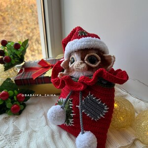 Christmas Gizmo amigurumi in Santa hat, placed among festive holiday decorations, handmade and eco-friendly gift idea.
