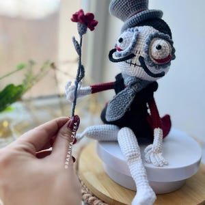 Crochet Mr. Santa Muerte in gray and black colors, holding a red flower, ideal for gothic or Halloween-inspired displays.