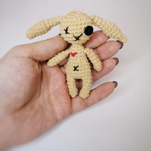 A small beige crochet voodoo bunny with black button eyes, an ‘X’ stitched on its belly, and long floppy ears, held in a person's hand.