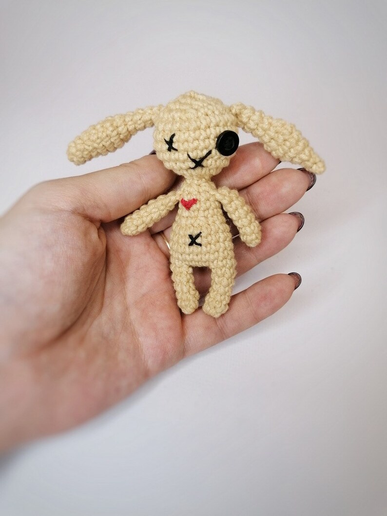 A small beige crochet voodoo bunny with black button eyes, an ‘X’ stitched on its belly, and long floppy ears, held in a person's hand.