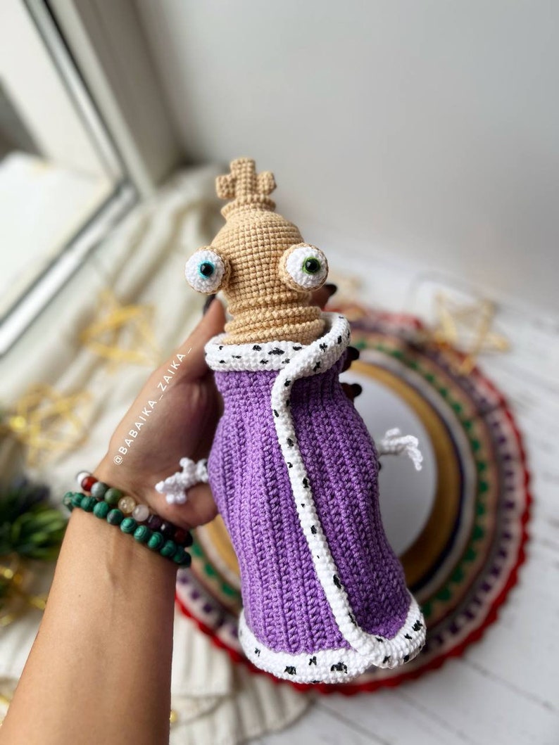 Close-up view of an intricately crocheted Kinger figure in a purple robe with a crown, set against a festive background.