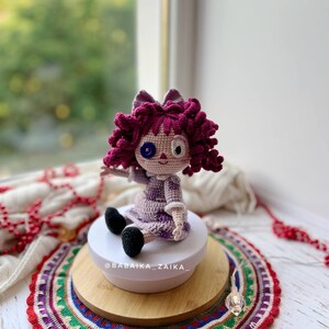 The crochet doll sitting on a white rotating stand, with red decorative beads in the background.