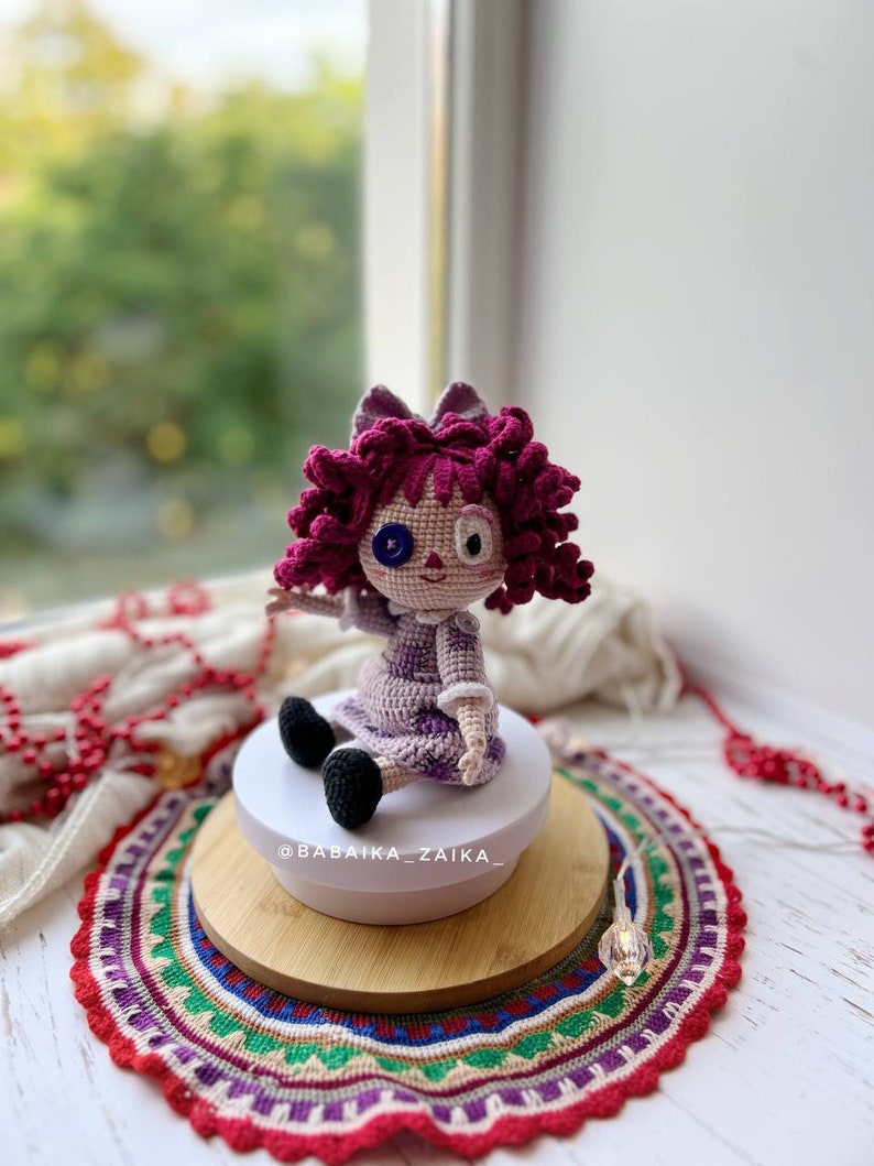 The crochet doll sitting on a white rotating stand, with red decorative beads in the background.